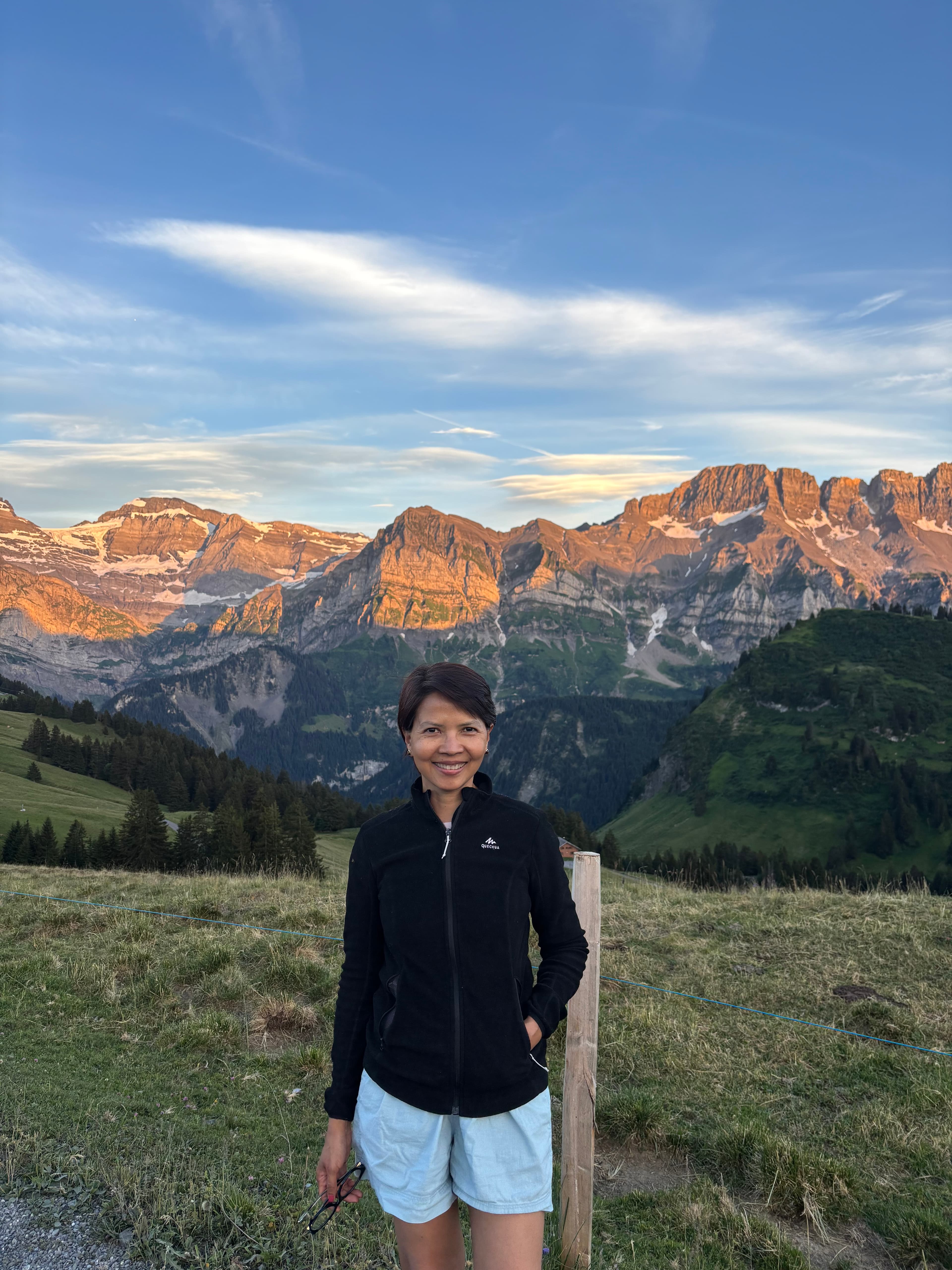 Smiling woman in a black jacket stands before golden mountain peaks under a blue sky.